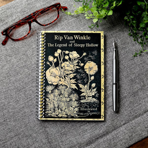 A travel-sized spiral notebook featuring a vintage book cover: Rip Van Winkle and The Legend of Sleepy Hollow by Washington Irving. Features a bold floral design. The colors are black and gold.