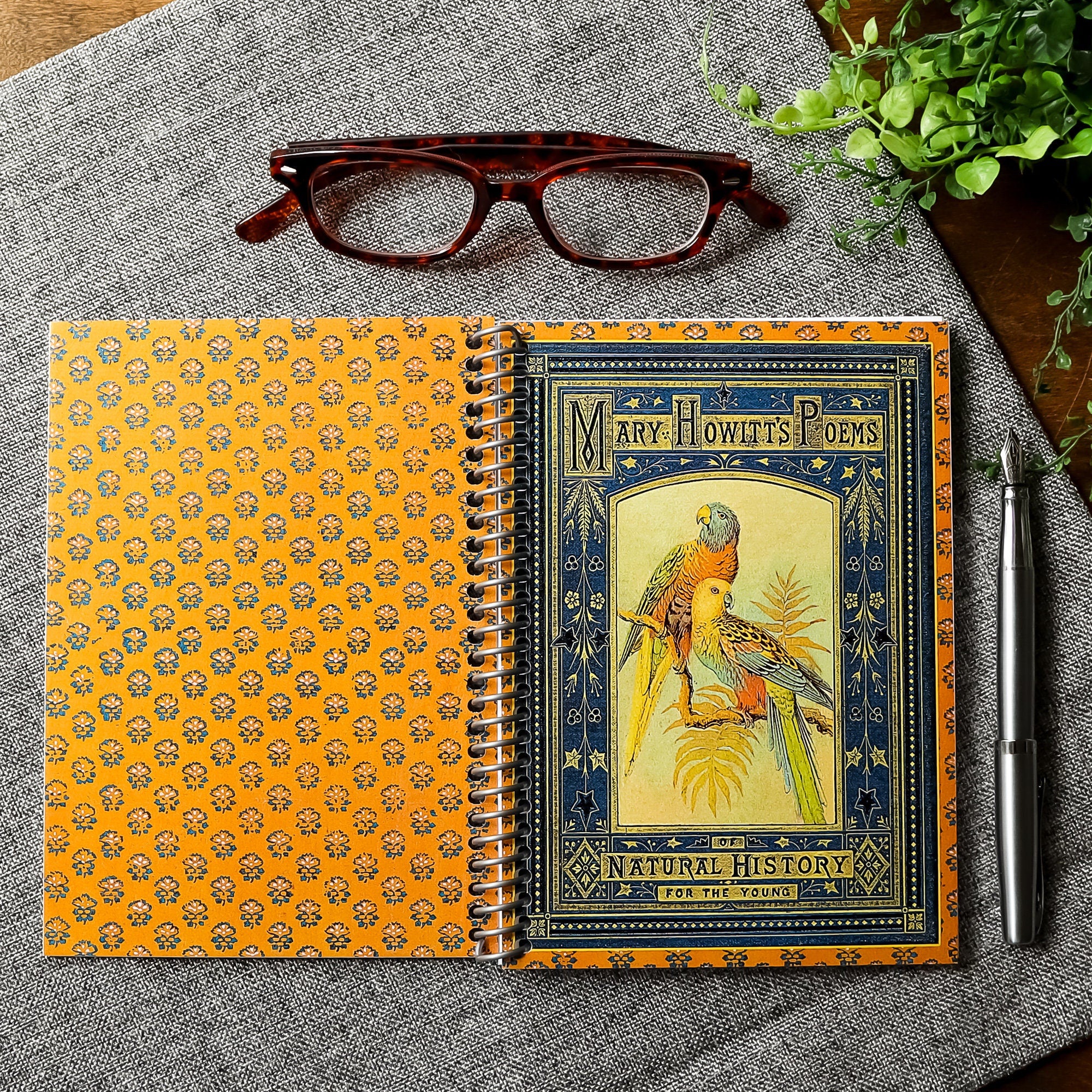 A travel-sized notebook featuring a vintage book cover: Mary Howitt&#39;s Poems of Natural History for the Young. Features a framed illustration of two colorful parrots. Colors are deep blue, orange, gold and green.