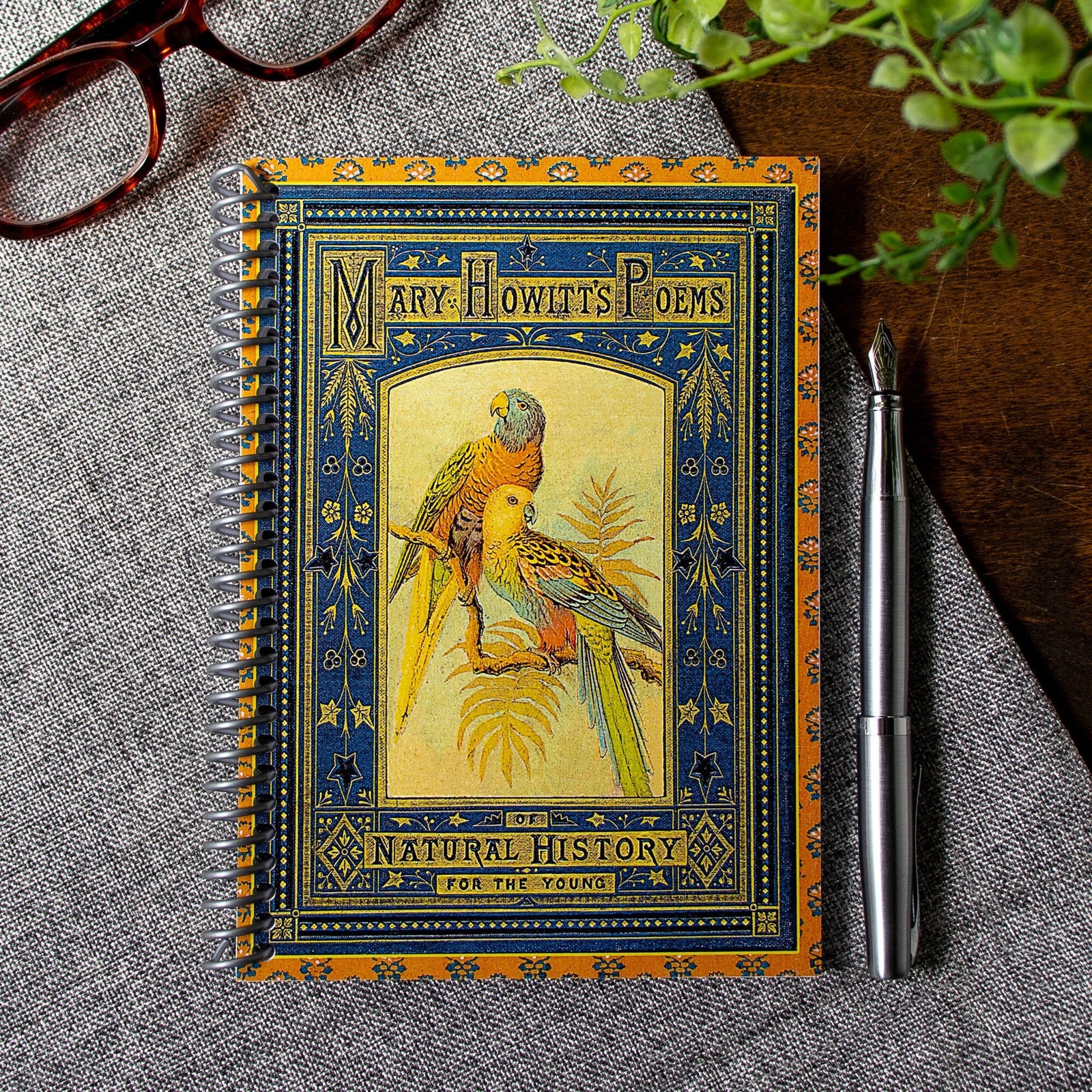 A travel-sized notebook featuring a vintage book cover: Mary Howitt's Poems of Natural History for the Young. Features a framed illustration of two colorful parrots. Colors are deep blue, orange, gold and green.