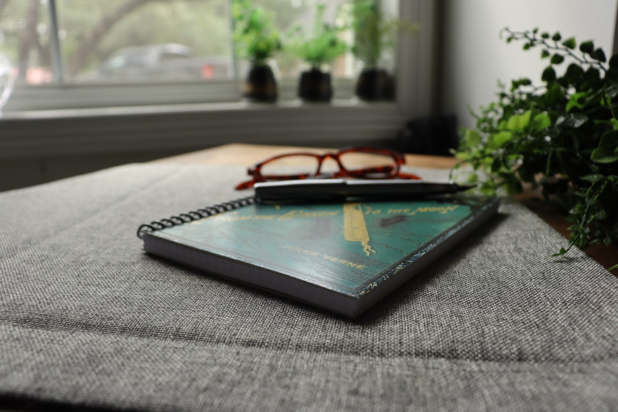 Detail image of a travel-sized spiral notebook featuring a vintage book cover: From the Earth to the Moon by Jules Verne. Features an illustration of a rocket ship flying towards the moon. The colors are deep green, blue, black, and gold.