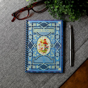 A travel-sized spiral notebook featuring a vintage book cover: Flora Symbolica or The Language and Sentiment of Flowers. Features a floral illustration surrounded by an ornate frame. The colors are sky blue, black, red, pink and gold.