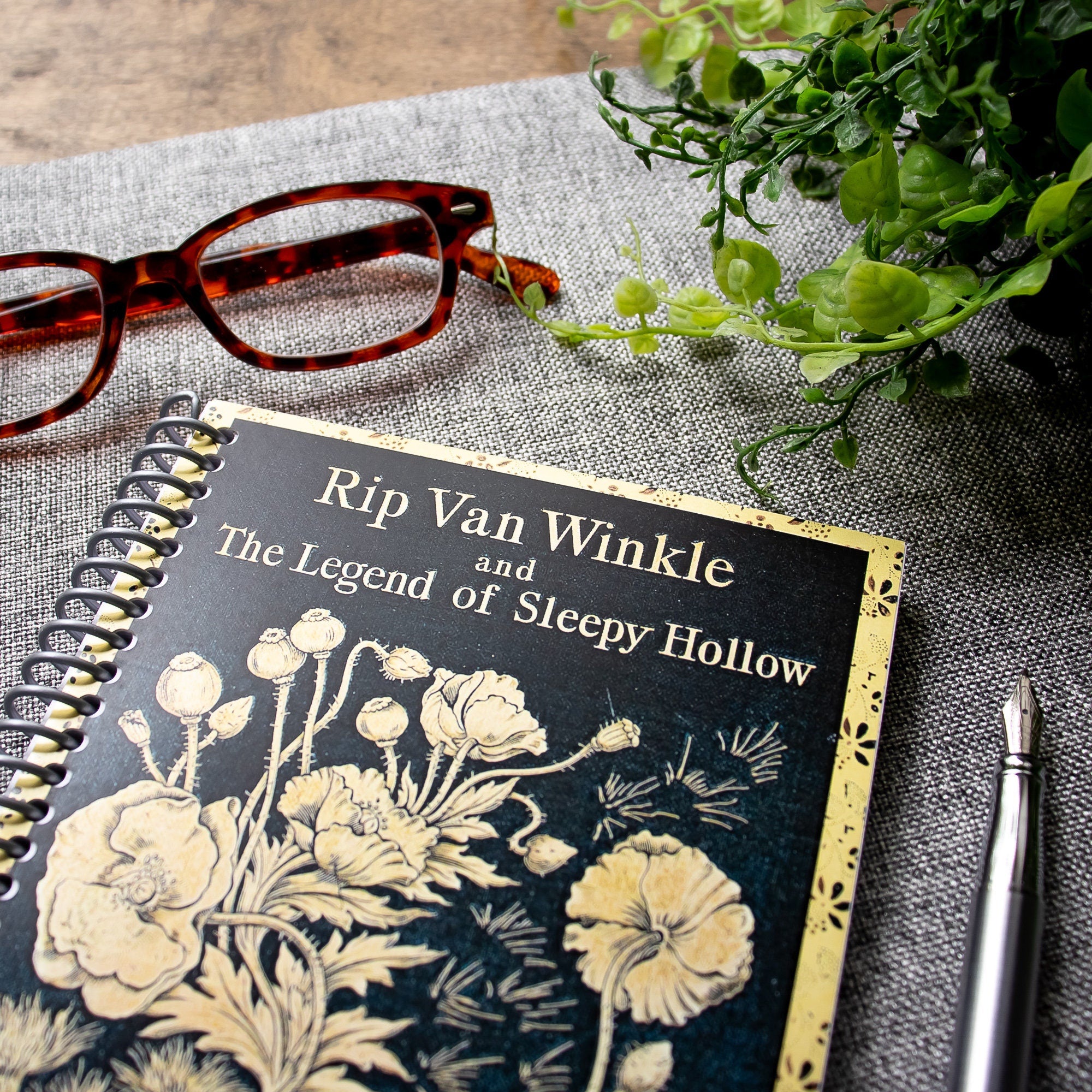 A travel-sized spiral notebook featuring a vintage book cover: Rip Van Winkle and The Legend of Sleepy Hollow by Washington Irving. Features a bold floral design. The colors are black and gold.