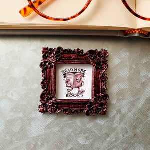 This is a small framed refrigerator magnet on a galvanized board, It features the phrase "Read More Books", and is made by SunshineandRavioli. The resin frame is ornate vintage style with a metallic burgundy finish. It measures 2.25 x 2.25 inches.