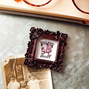 This is a small framed refrigerator magnet holding a photo on a galvanized board, It features the phrase "Read More Books", and is made by SunshineandRavioli. The resin frame is ornate vintage style with a metallic burgundy finish. It measures 2.25 x 2.25 inches.